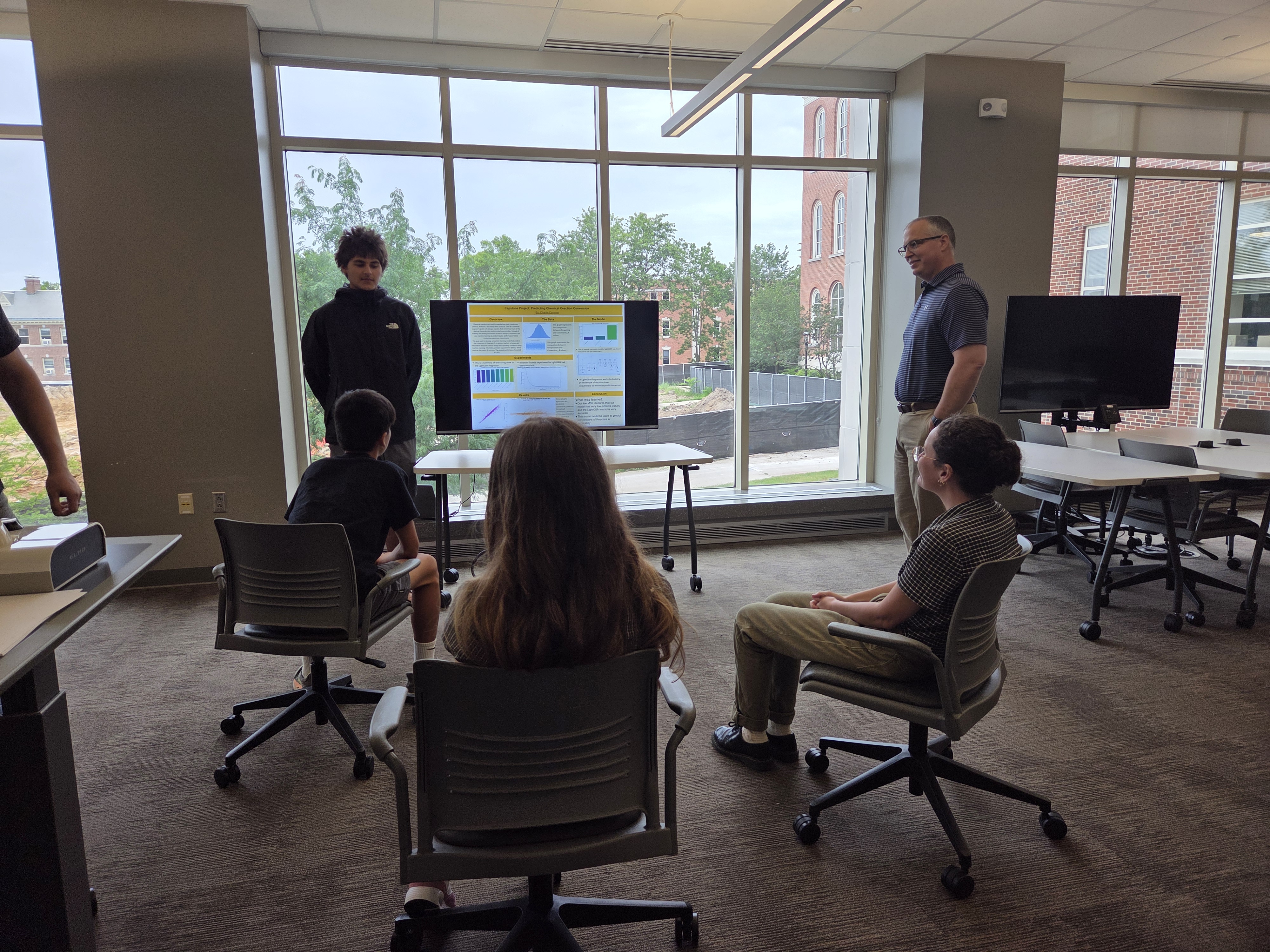 A camp participant presents a capstone poster on predicting chemical reaction conversion to instructors and fellow campers